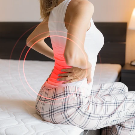 A woman sitting on a bed while holding her lower back in pain.